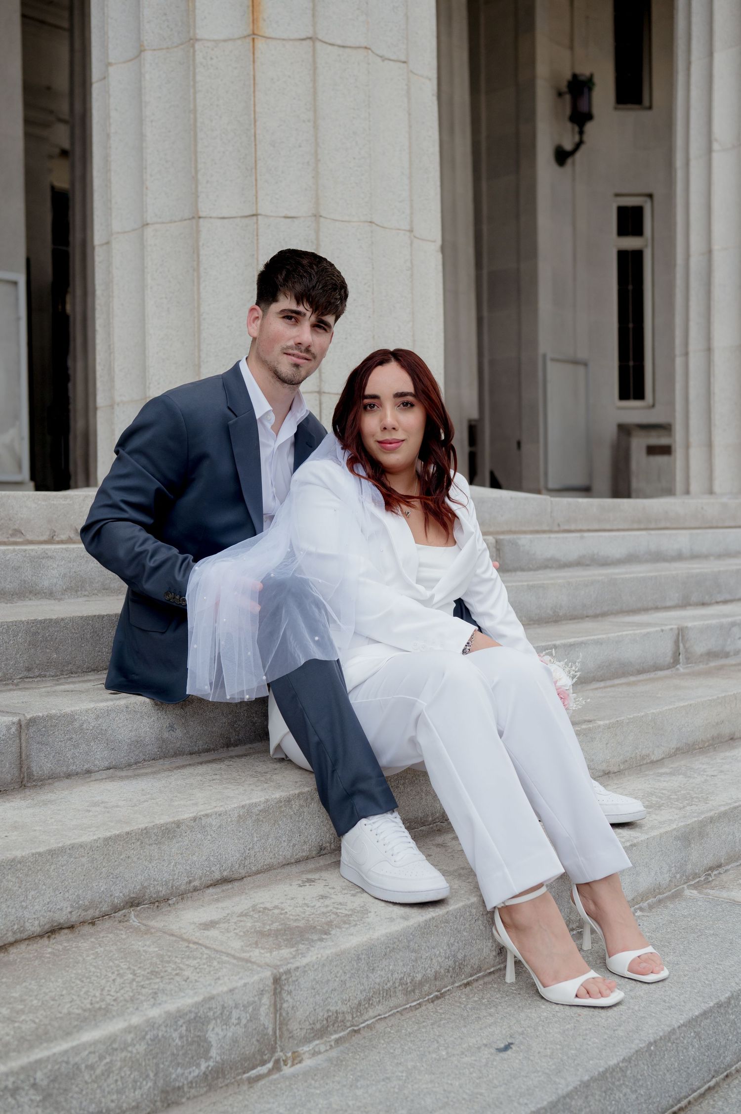 A couple in formal attire sitting together on white stone steps with architectural columns visible in the background.