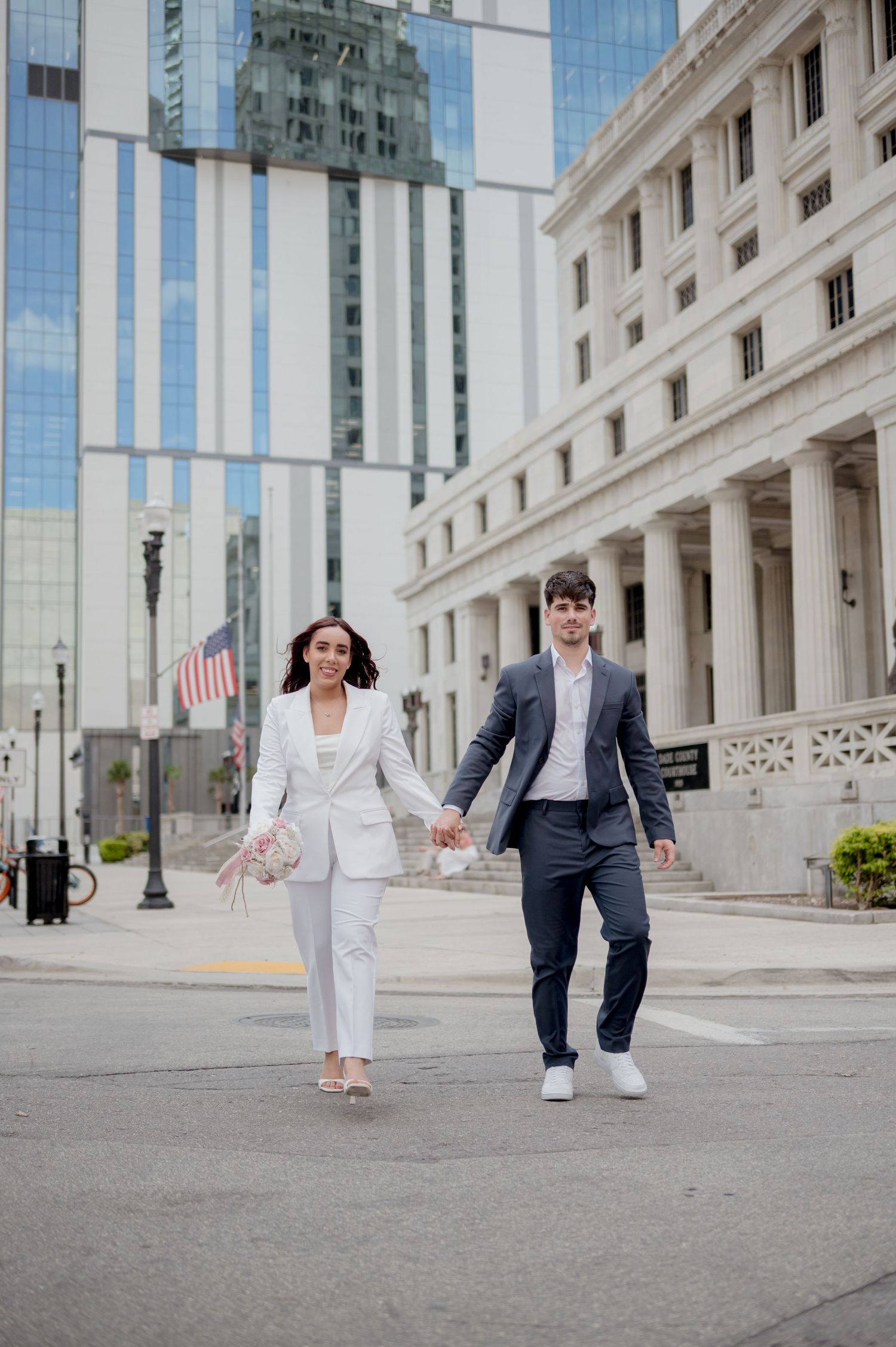 A couple in formal attire walking together playfully in front of a grand stone and glass building.