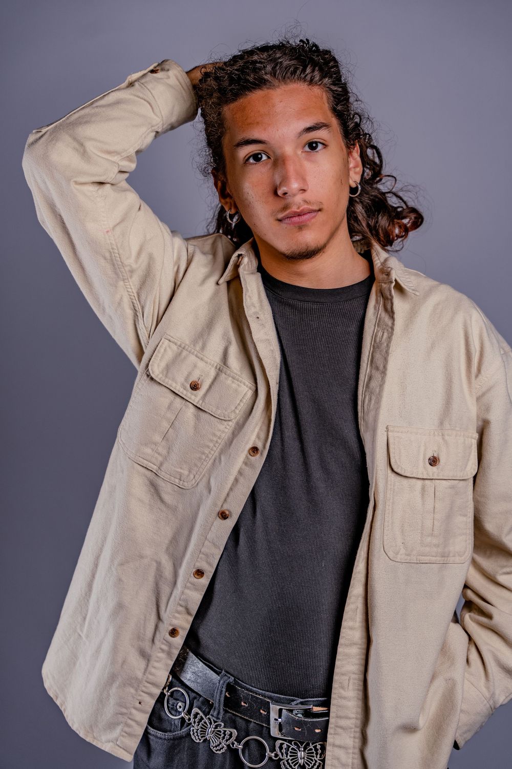 Model in beige utility jacket poses with hand on head against gray backdrop.
