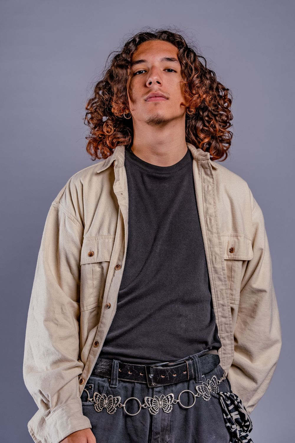 Fashion portrait of person with curly hair wearing beige jacket and chain belt.
