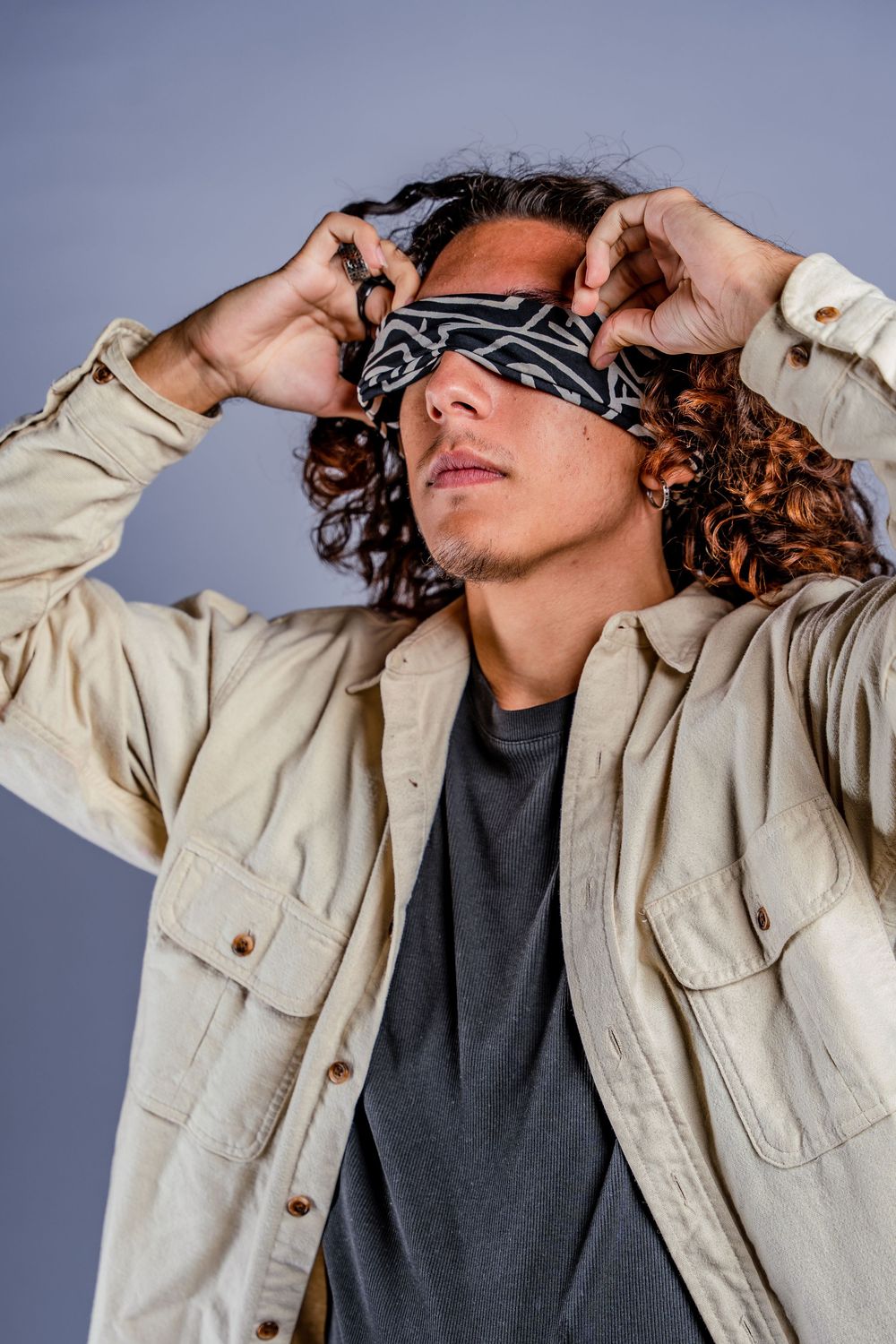 Model wearing beige jacket adjusts black bandana around eyes.