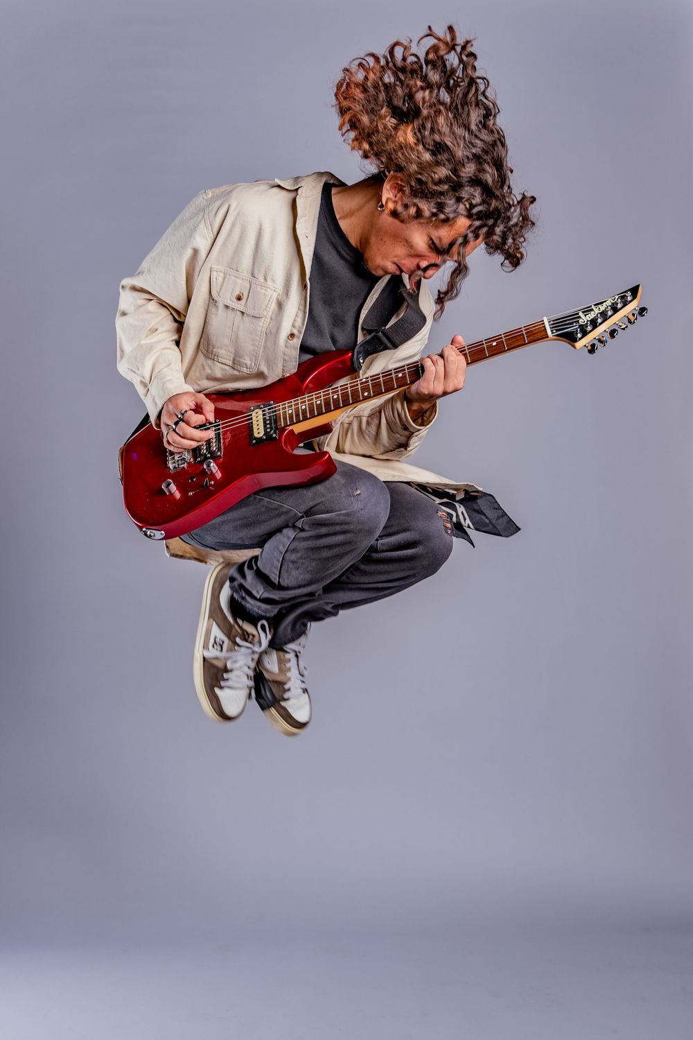 Musician with curly hair jumping while playing red electric guitar against gray background.