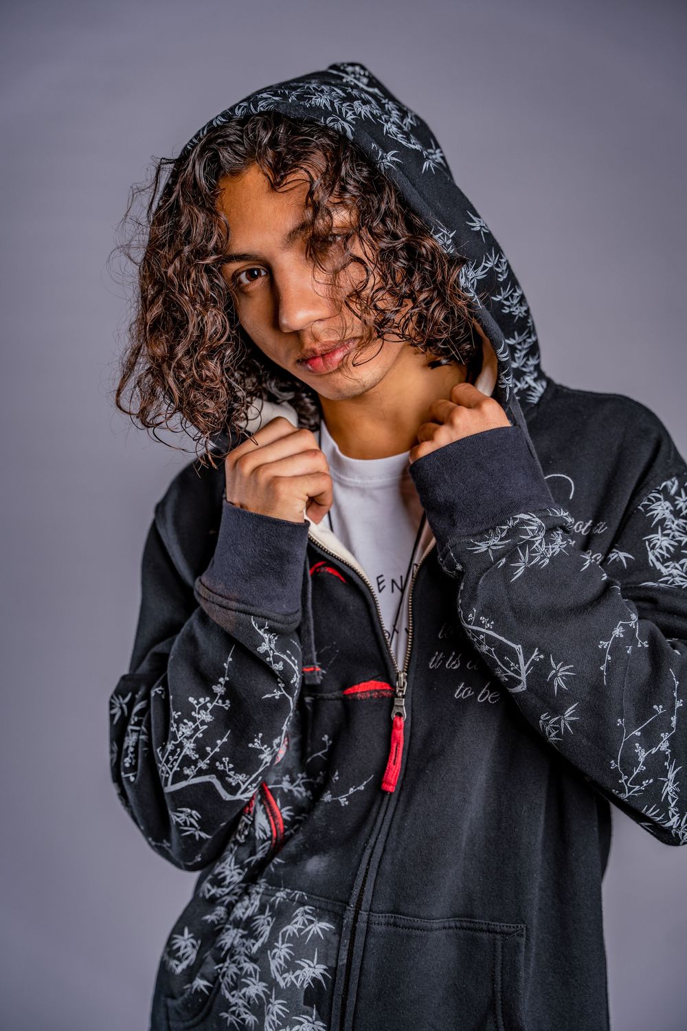 Person wearing black patterned hoodie poses against gray studio background.