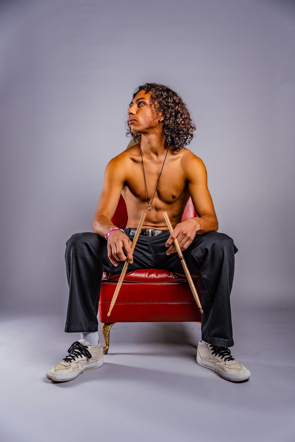 Shirtless figure sits casually on red chair wearing black pants and white sneakers.