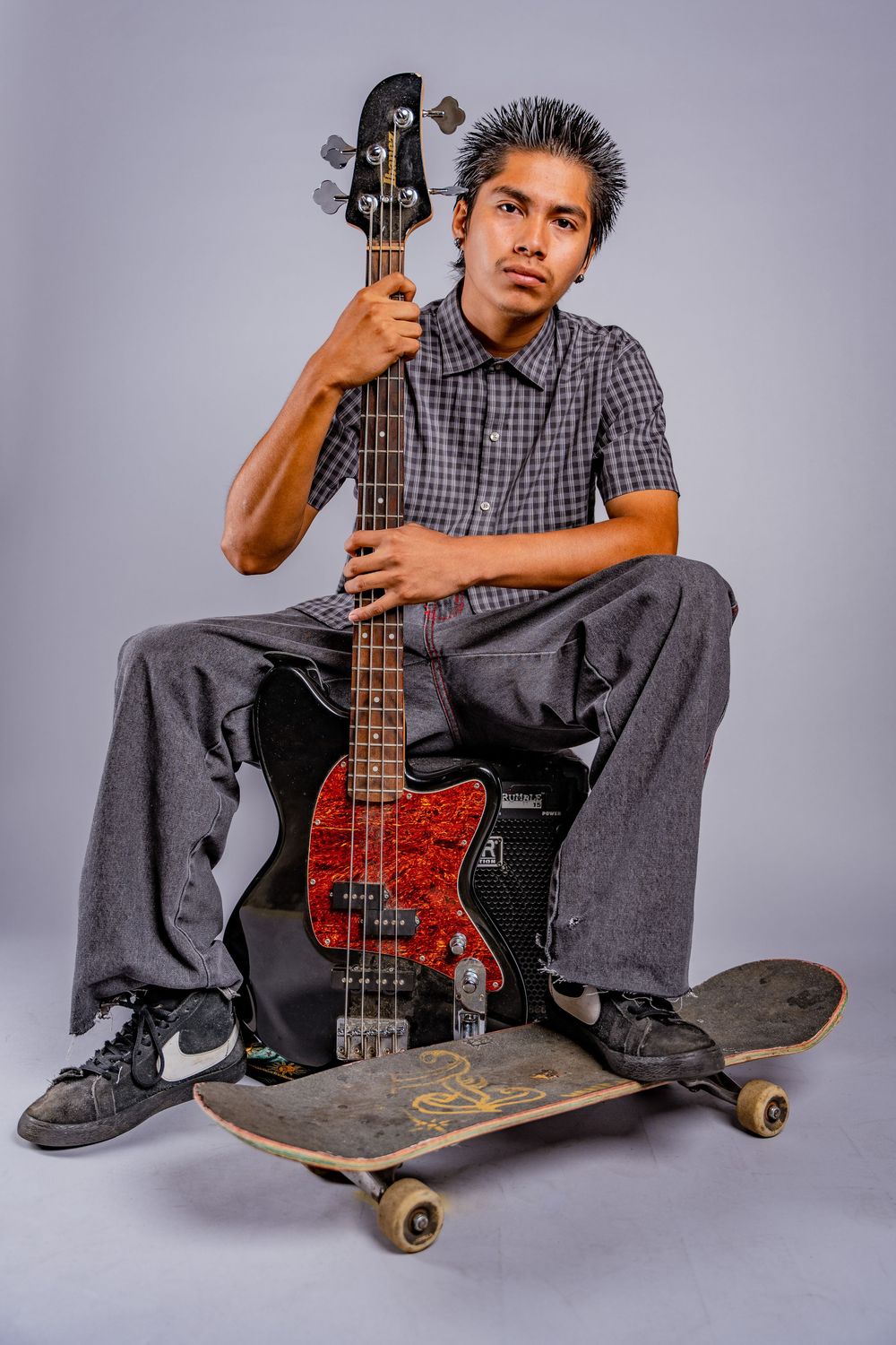 Skater sits on skateboard holding red and black bass guitar in casual studio setting.