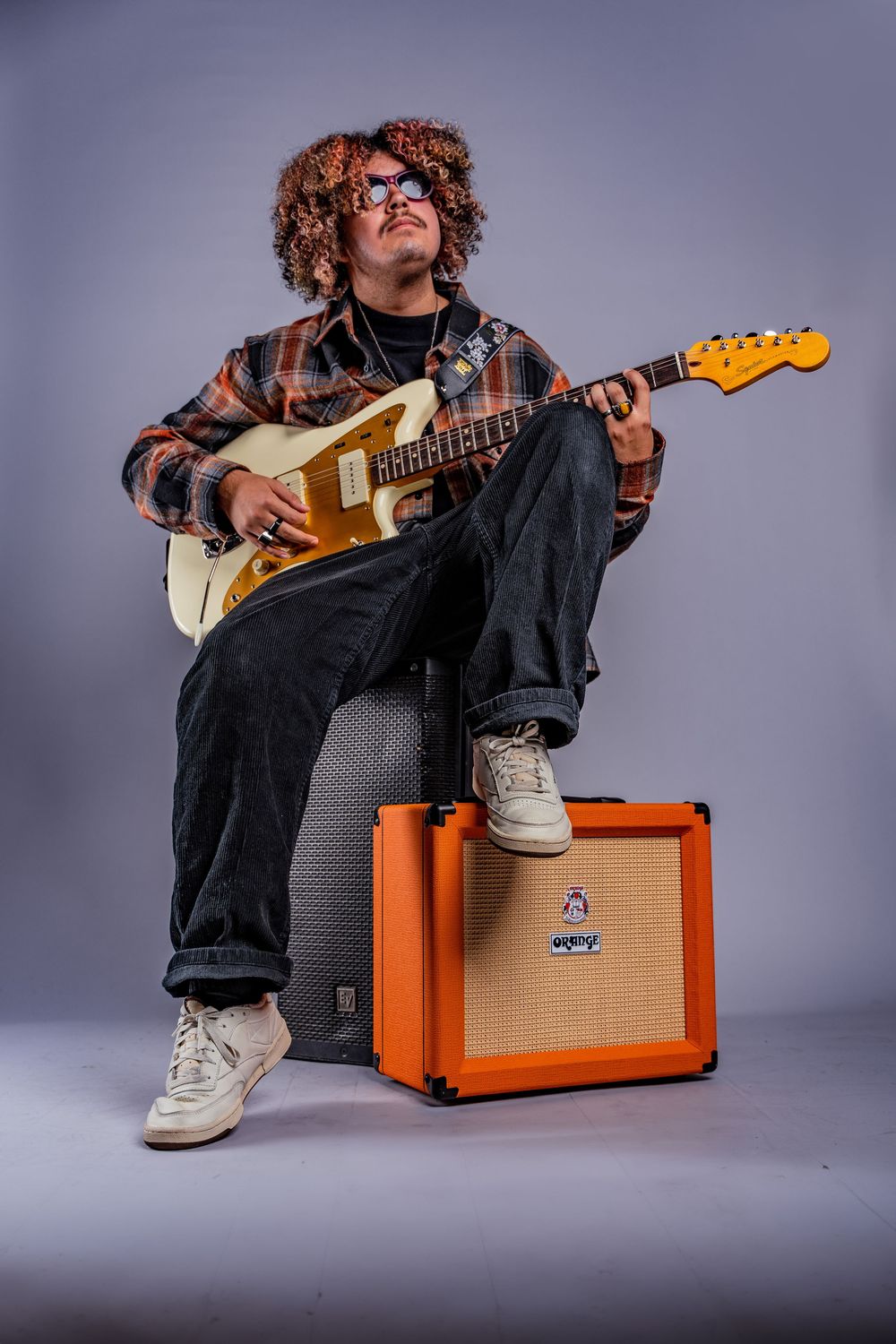 Musician sits with electric guitar next to orange amplifier in studio setting.