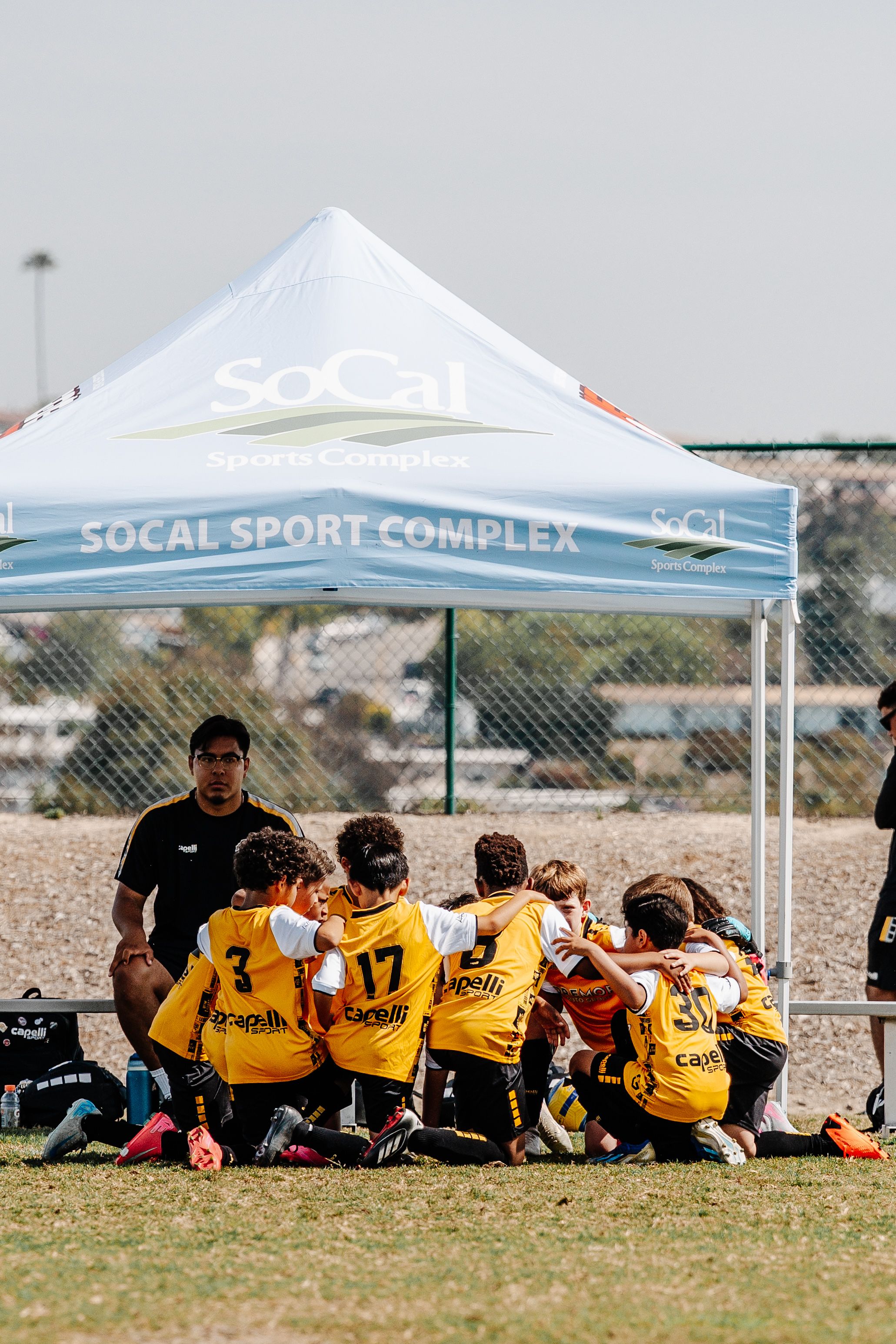 Youth soccer team huddles under white SoCal Sport Complex tent during match.
