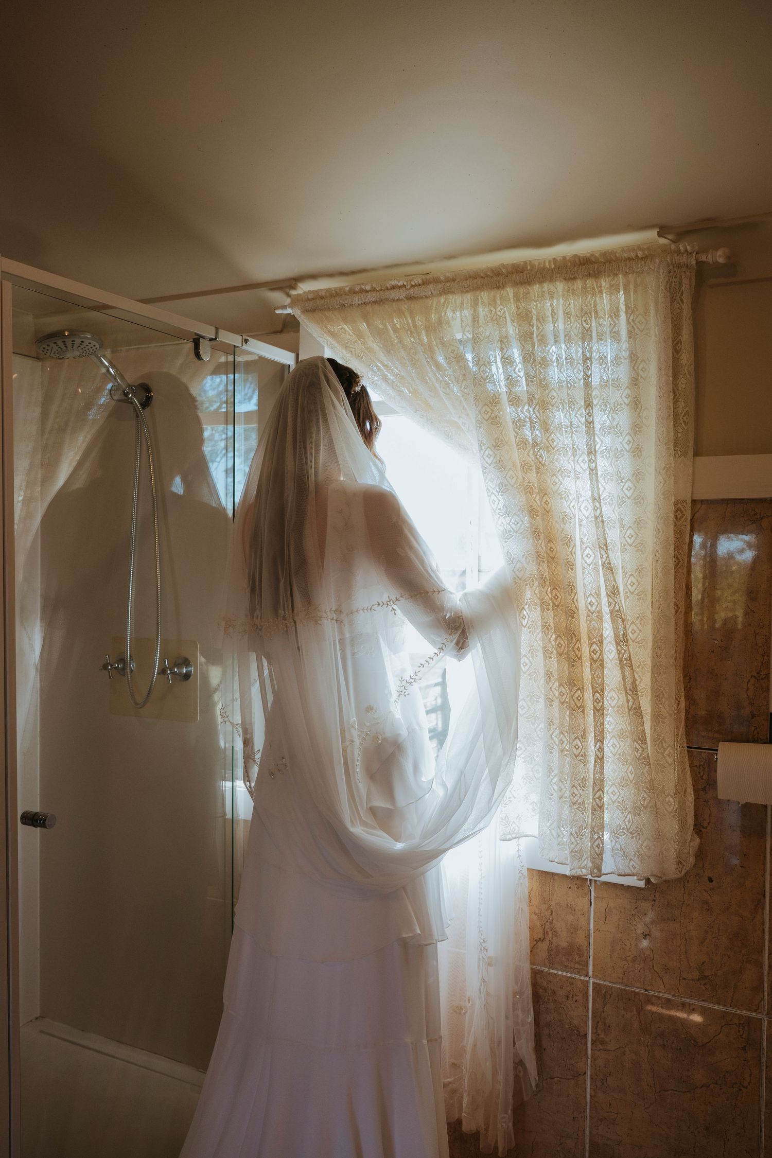 Wedding dress hanging in bathroom with sunlight streaming through sheer curtains.