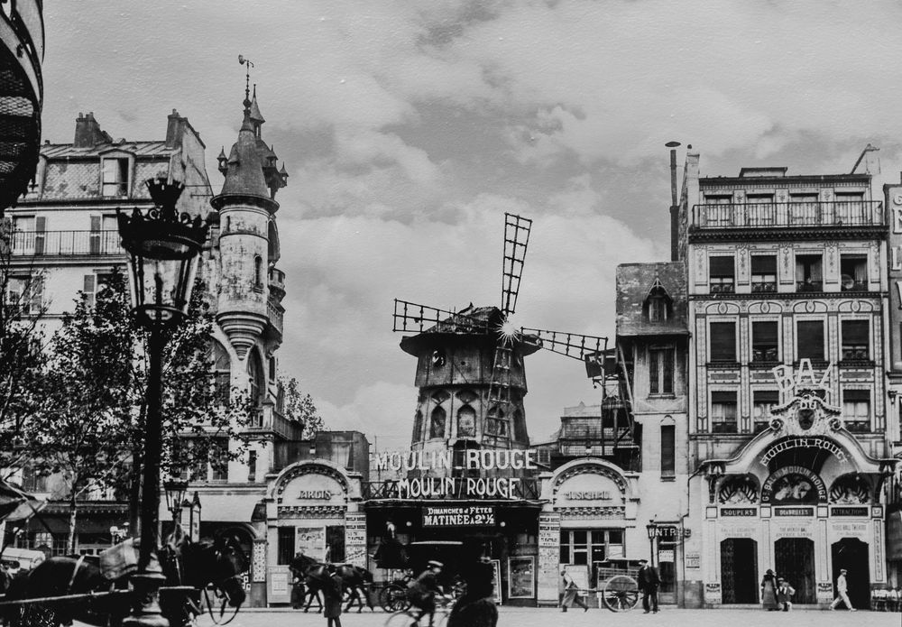 A black and white historical photograph shows a busy urban street scene with storefronts and cars from the early 1900s.