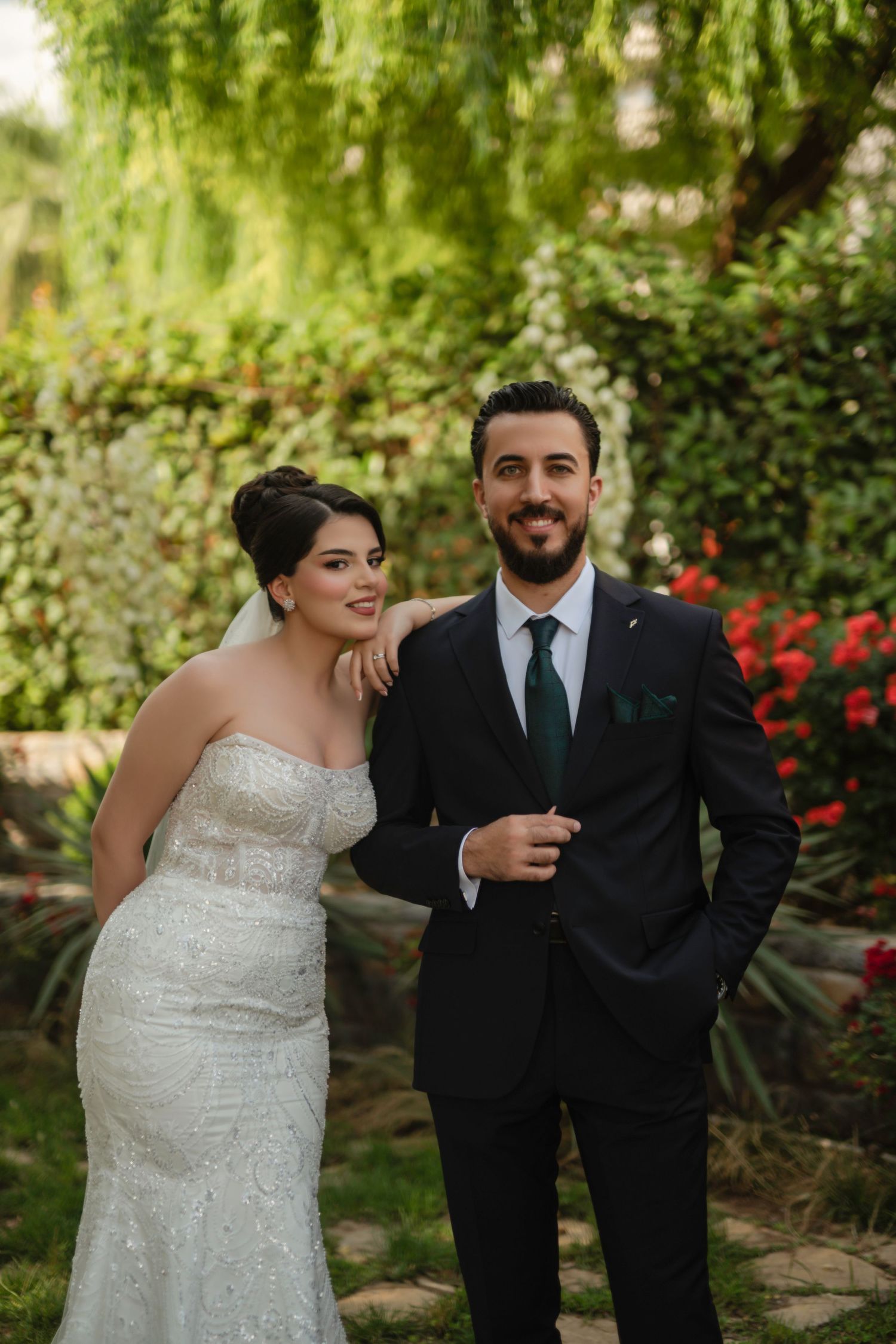 A couple poses together in a garden setting with willow trees and red flowers in the background.
