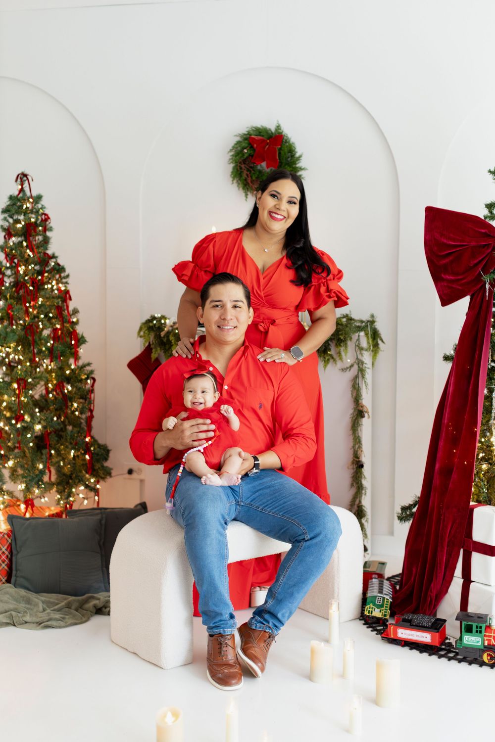 A family poses for Christmas photos in matching red outfits in front of a decorated tree and white backdrop.