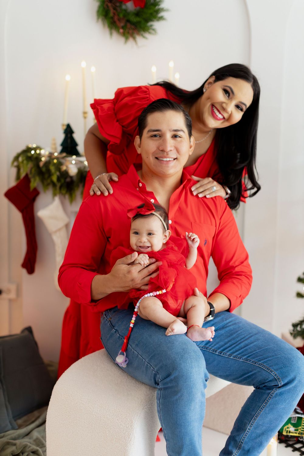 Family wearing matching red tops and jeans poses together for Christmas photos with holiday decor in background.
