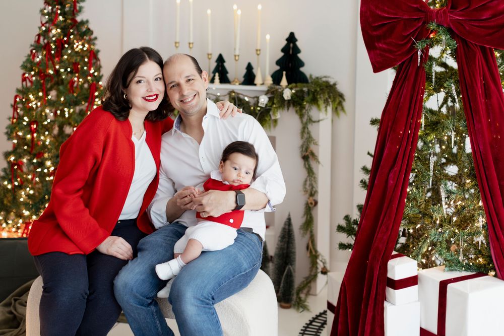Family poses together in front of Christmas trees and red curtains wearing coordinated red and white holiday outfits.