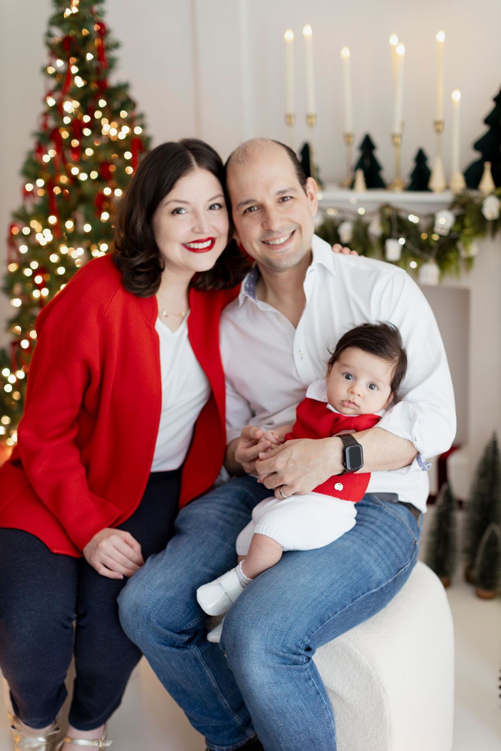 A family poses together in festive red and white outfits in front of a decorated Christmas tree and lit candles.