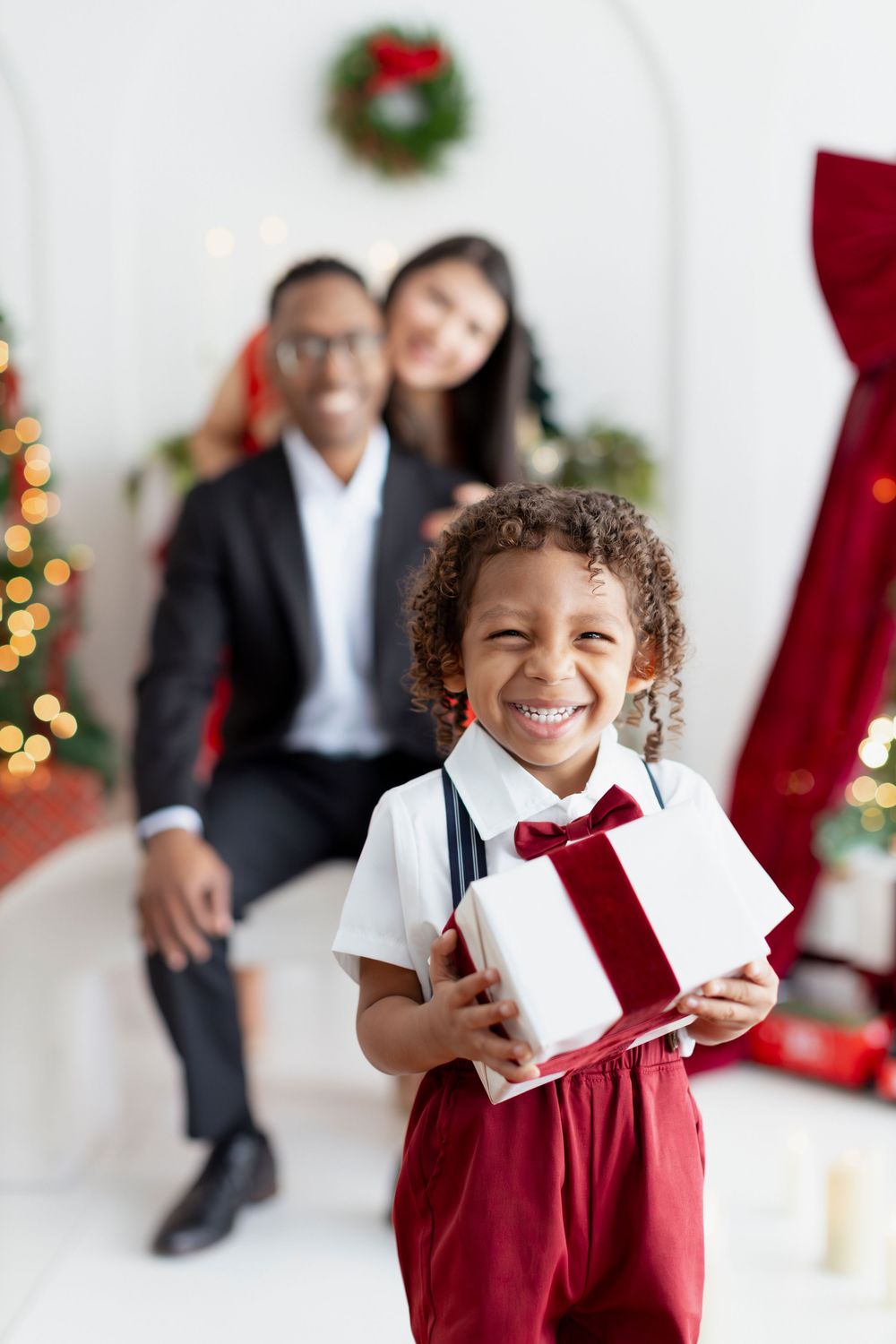 A child in red suspenders and bow tie holds a white Christmas gift box while standing in front of holiday decorations.