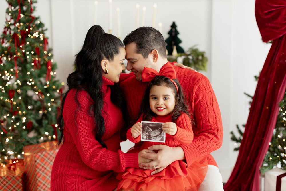 Family in matching red sweaters share intimate holiday moment by Christmas tree while child holds ultrasound photo.