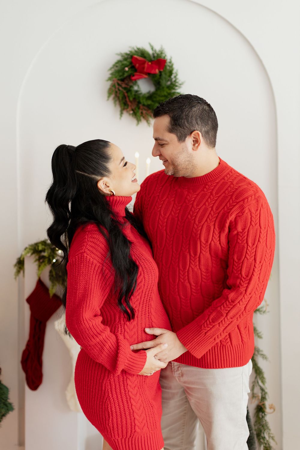 Couple in matching red sweaters share intimate moments against a white wall with holiday decorations.