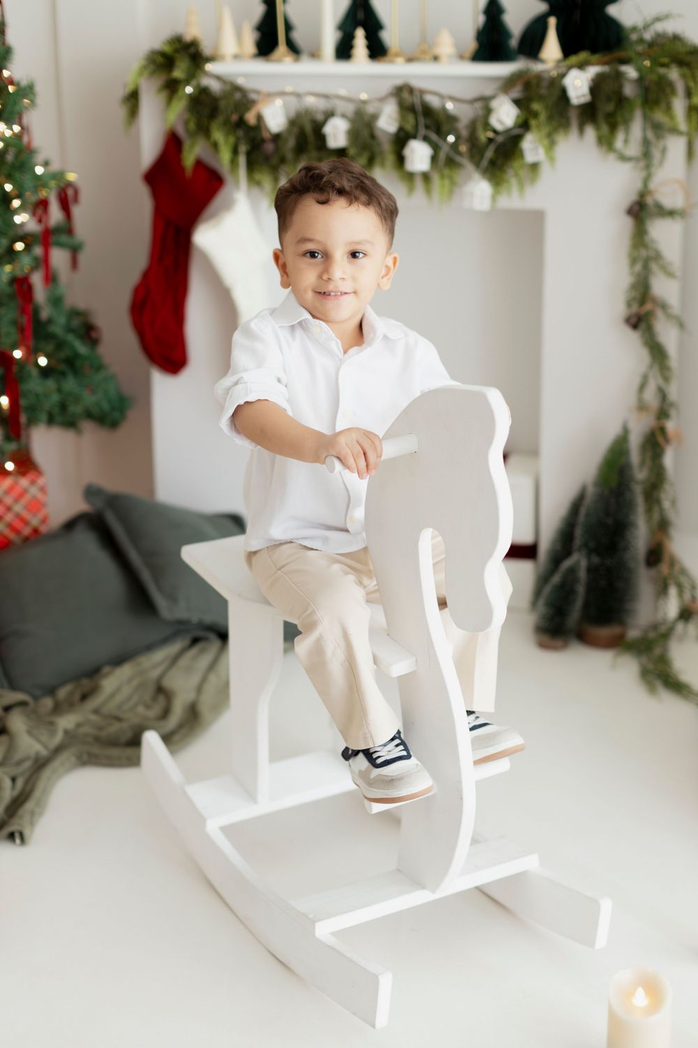 Child in white clothing sits on rocking horse with Christmas decorations and stockings hanging in background.