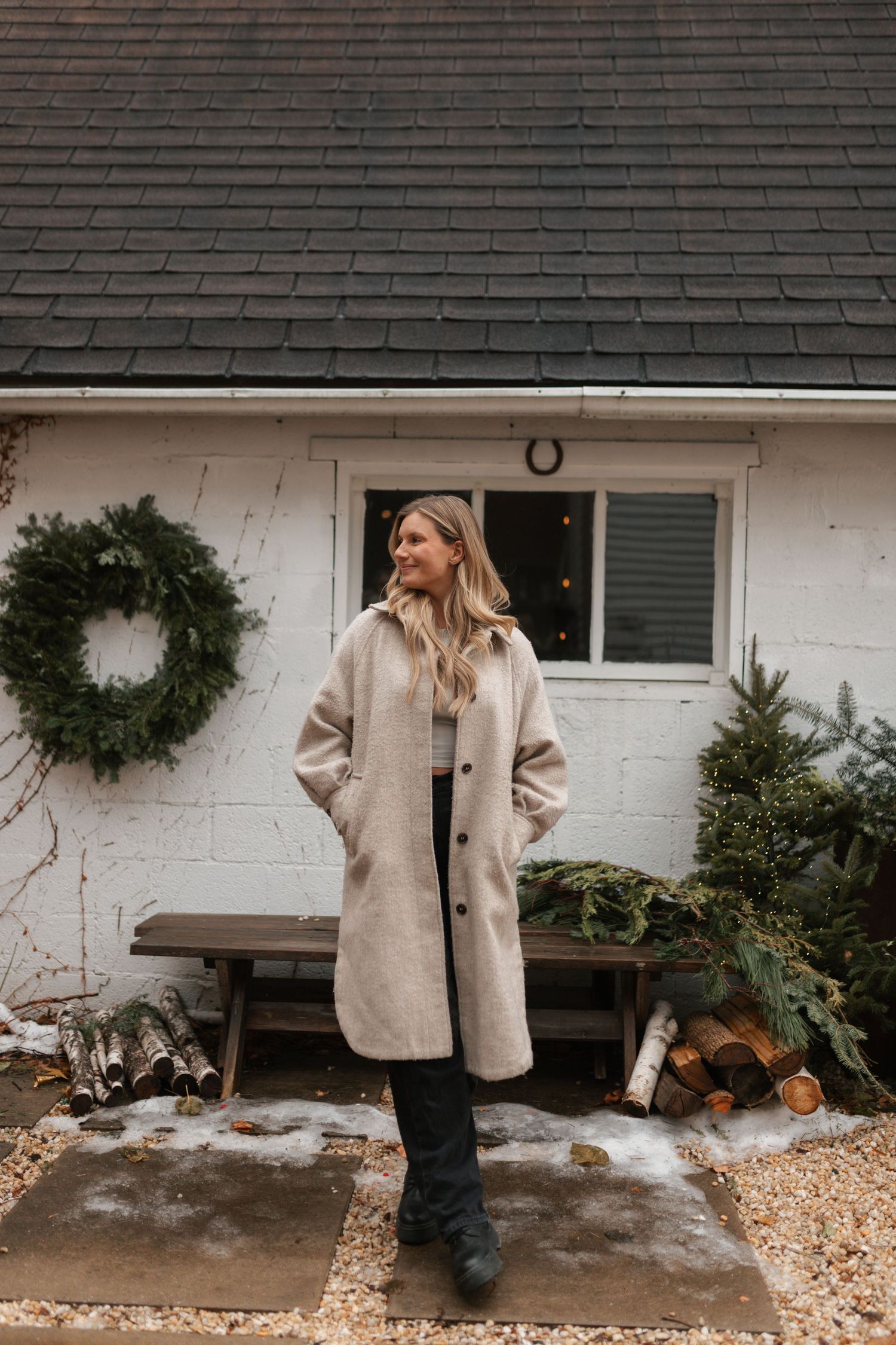 Smiling blonde woman in beige wool coat standing outside white cottage with Christmas wreath in winter.