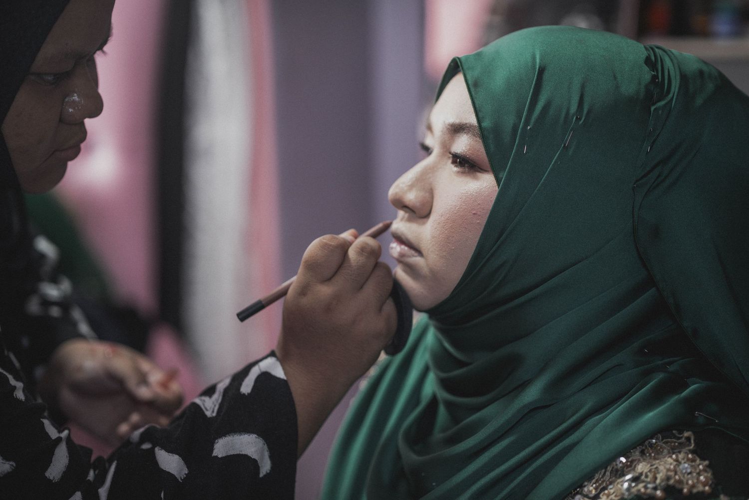 Makeup artist applying cosmetics to someone wearing a green hijab in soft lighting.