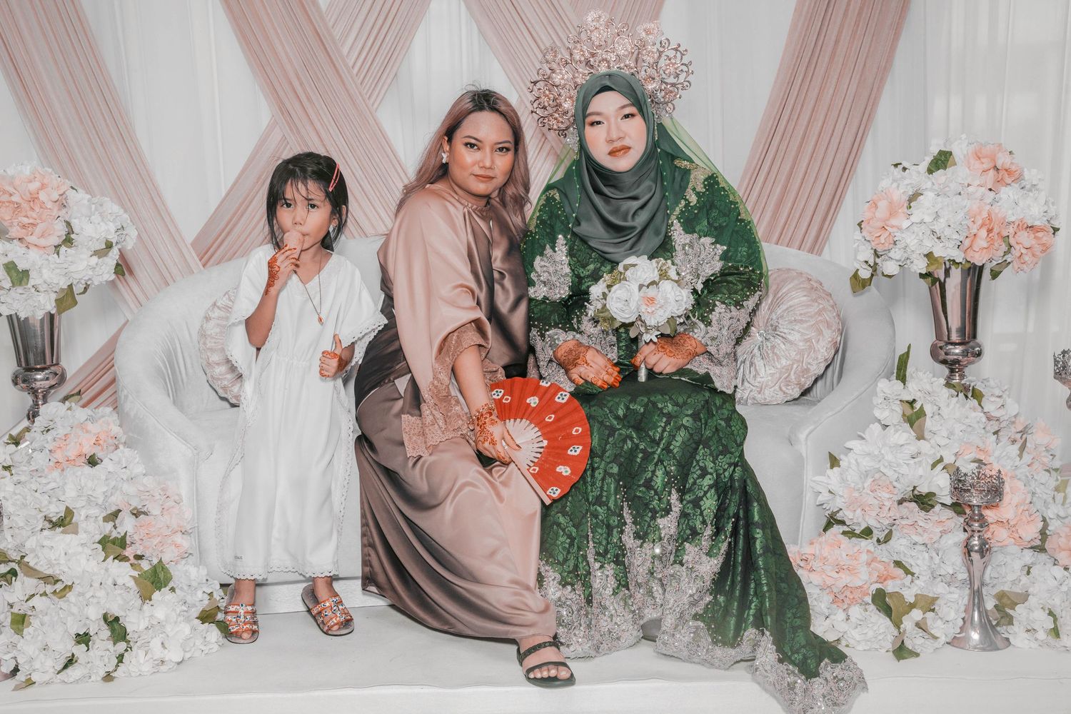 Three people pose in formal attire against pink and white floral backdrop at wedding celebration.