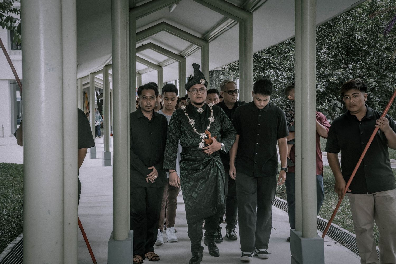 Group walks through covered walkway wearing formal black attire during wedding procession.