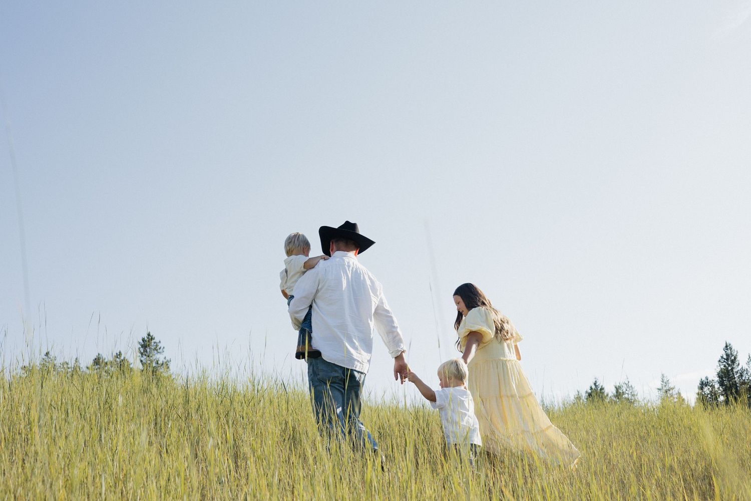 Black and white photo of a tender family moment outdoors in a field with pine trees in the background.