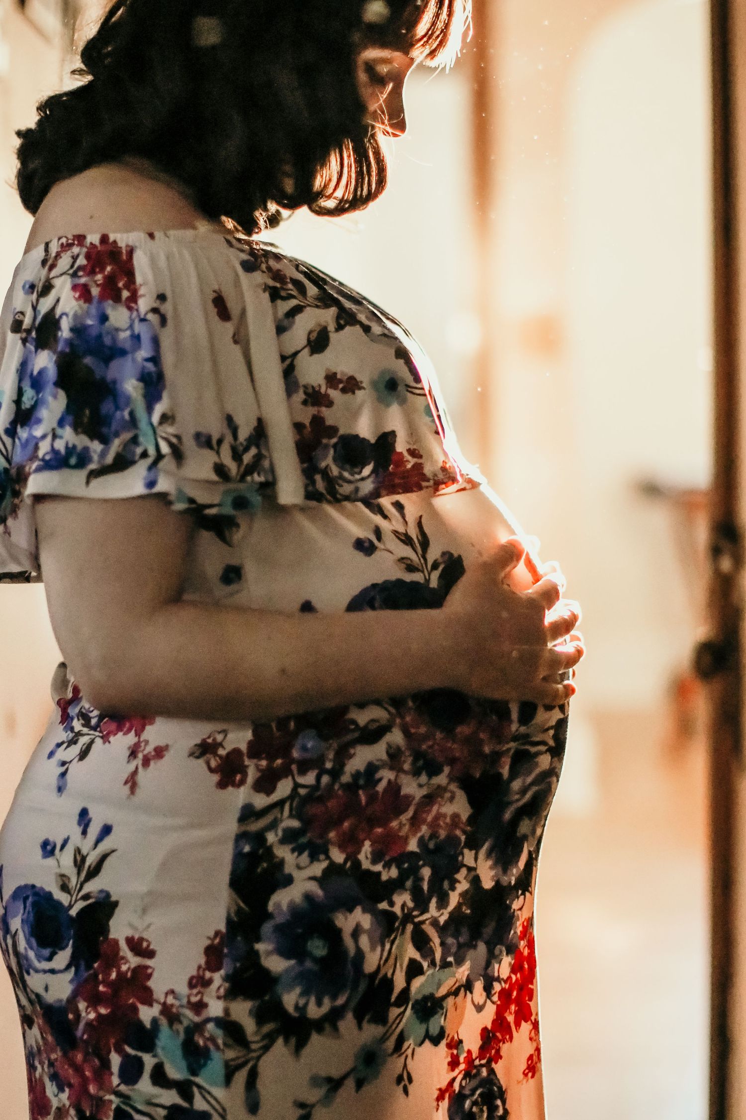 Side view of a pregnant woman in a floral dress standing in soft natural light.