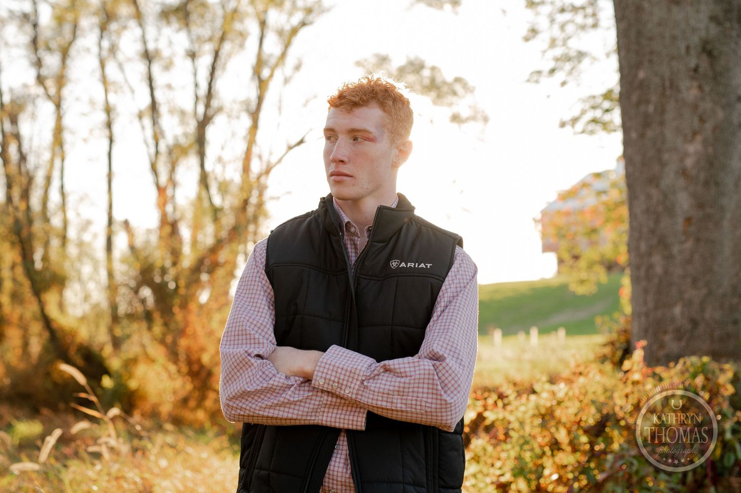 Person in black vest and plaid shirt standing with crossed arms in golden autumn setting.