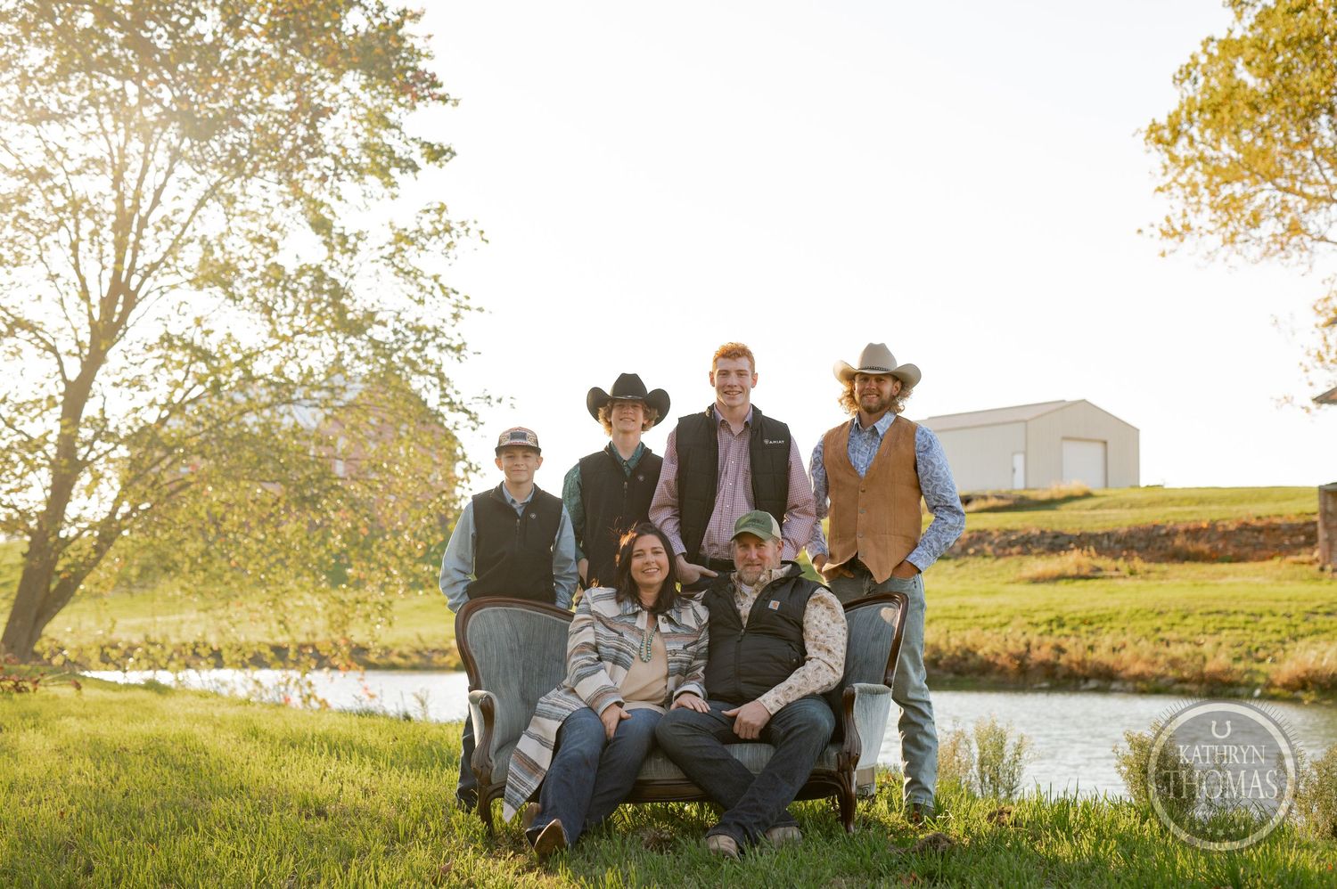 Family group portrait at sunset by water with rustic building on hillside in background.