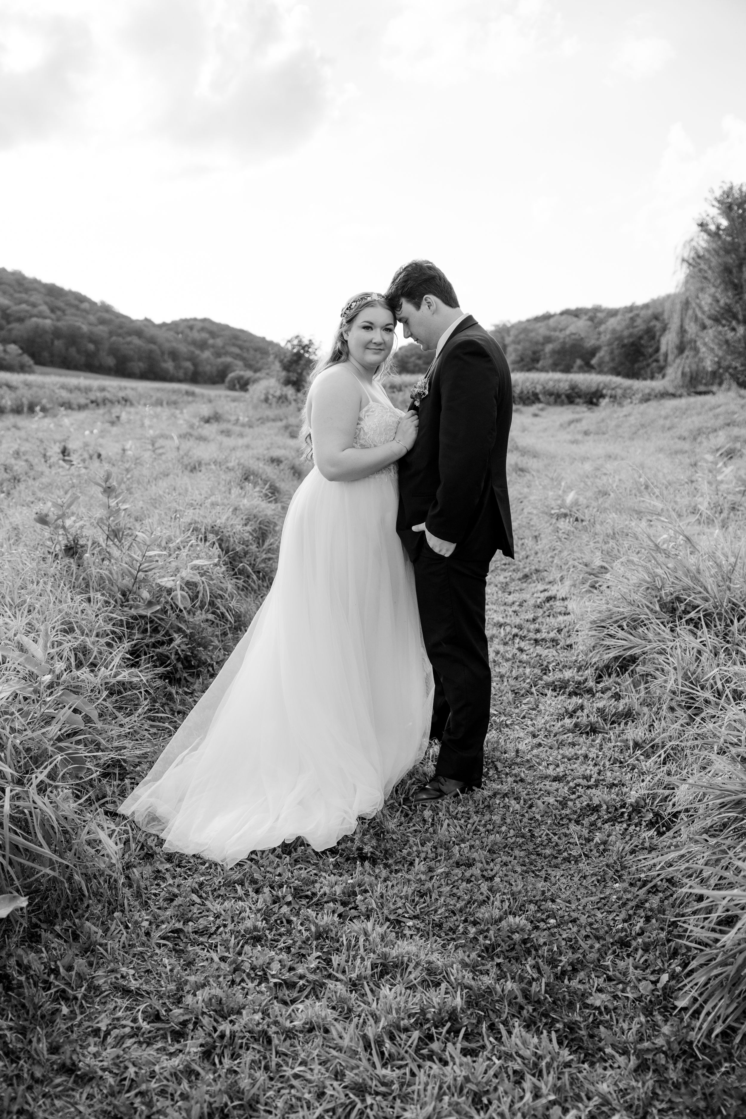 A romantic black and white wedding portrait of a couple standing together in a scenic field.
