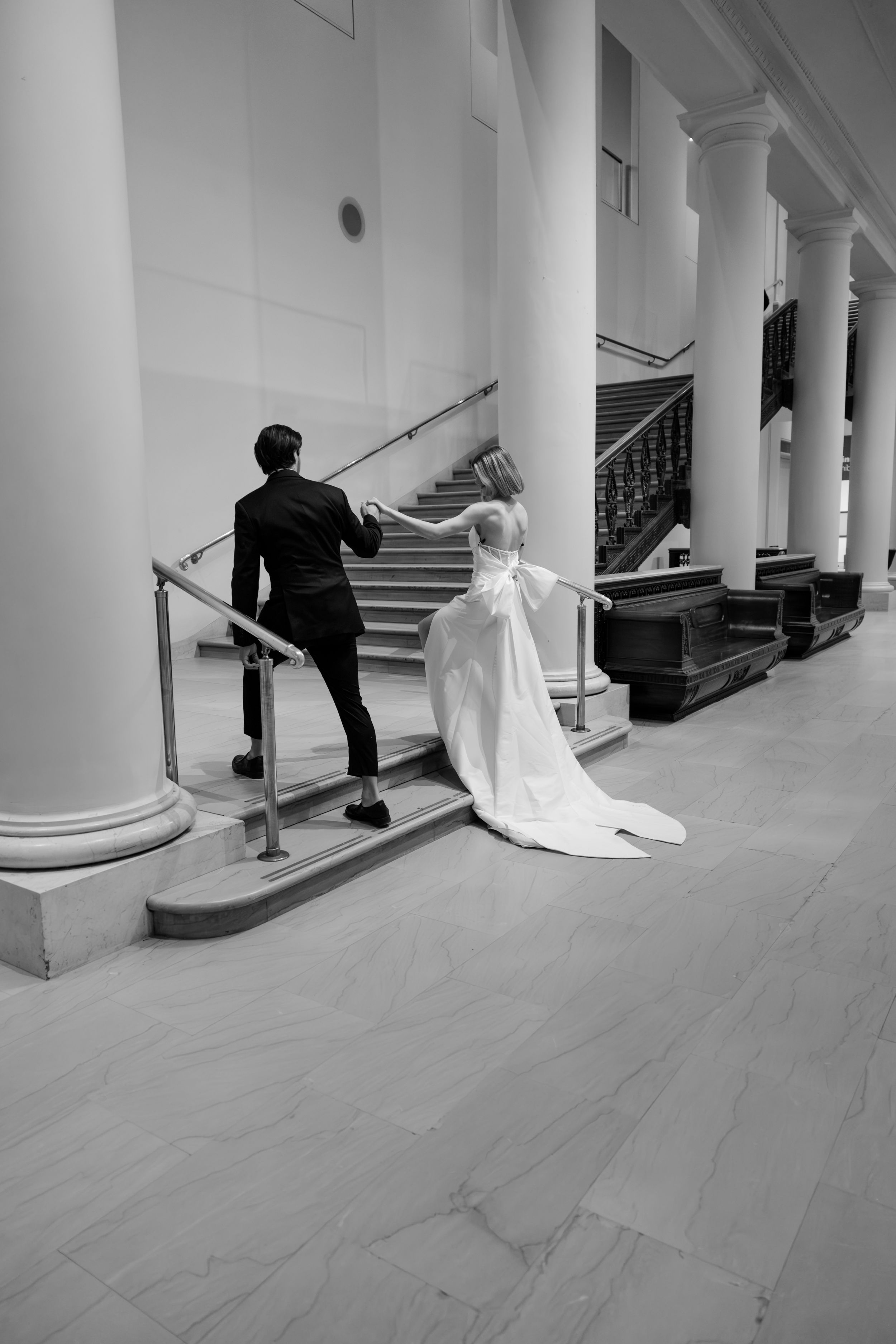 Black and white photo of a couple ascending marble stairs in formal wedding attire.