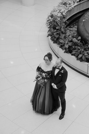 A couple in formal attire poses in an elegant indoor venue with a dramatic black wedding gown.