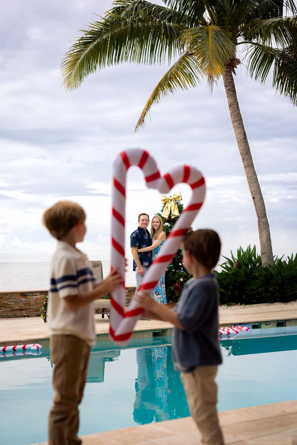 Children hold candy cane decorations by a poolside with palm tree and cloudy sky at a tropical holiday setting.