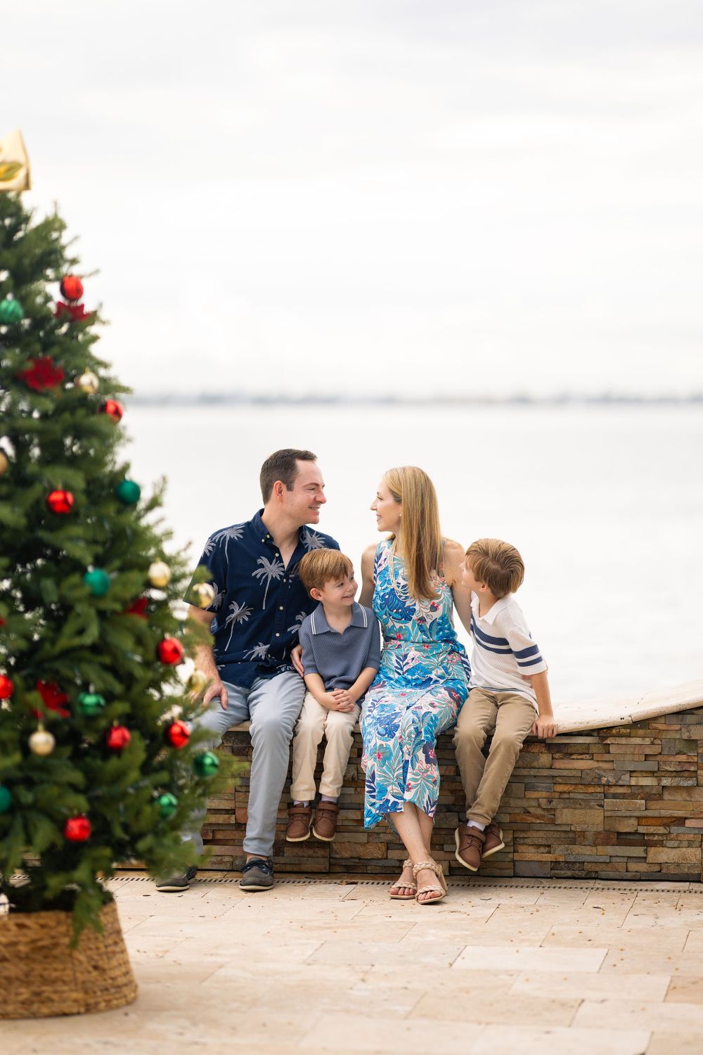 A family sits together on a stone wall next to a decorated Christmas tree during a holiday photo session.