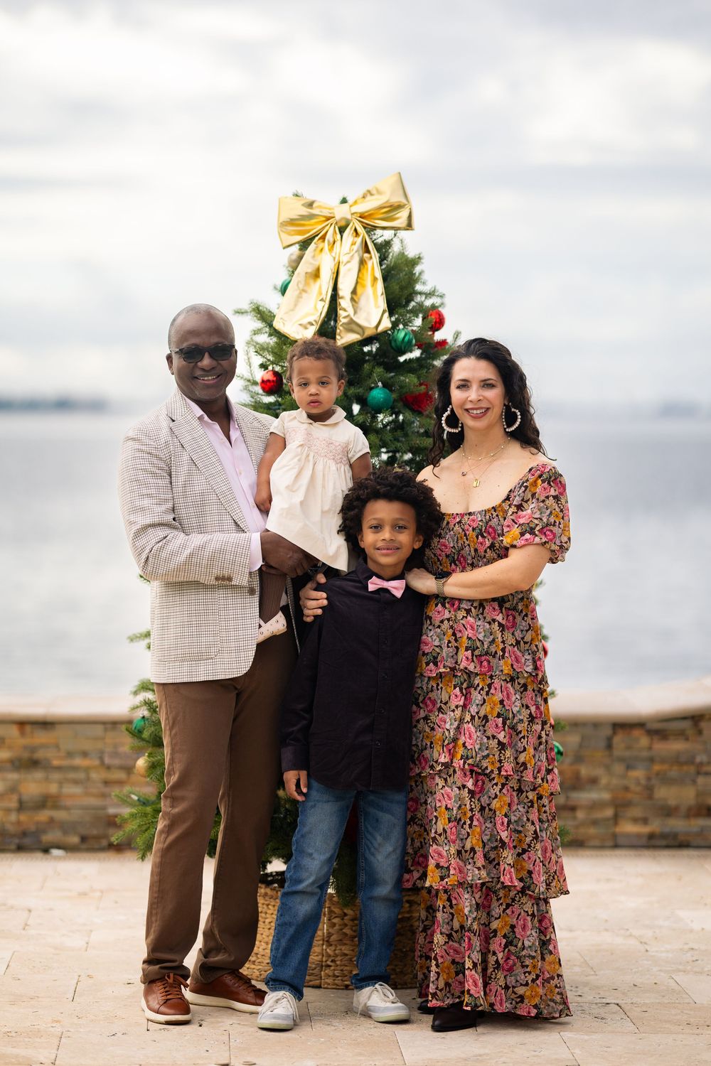A family poses together outdoors on a stone patio during a holiday photoshoot with a decorated Christmas tree.