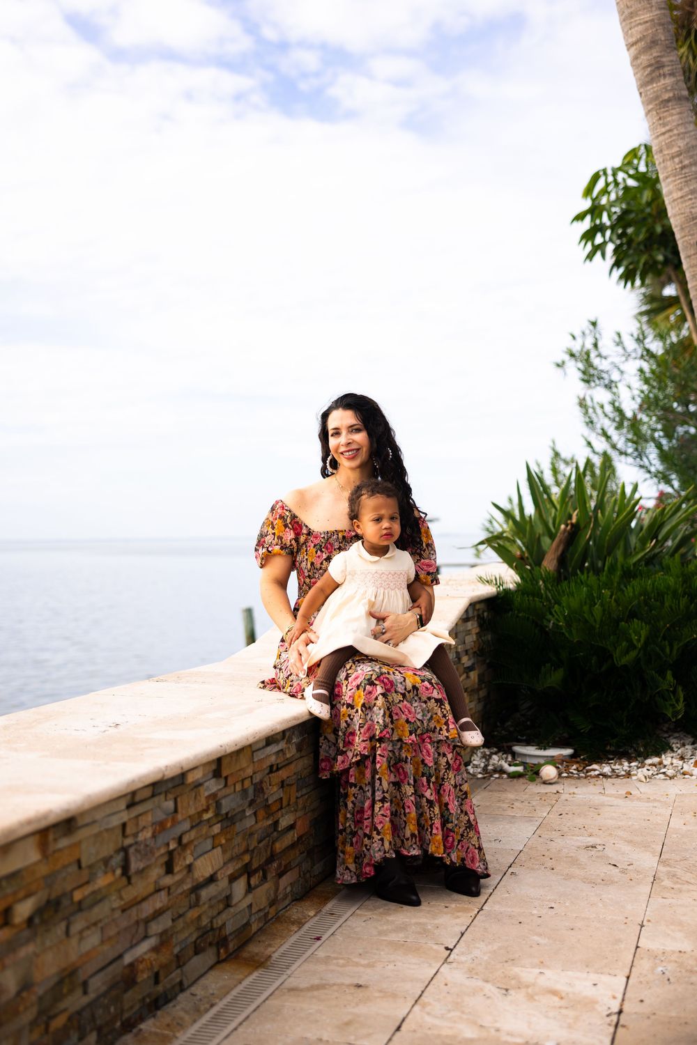 A woman in a floral dress sits on a stone wall overlooking the ocean on a sunny day.