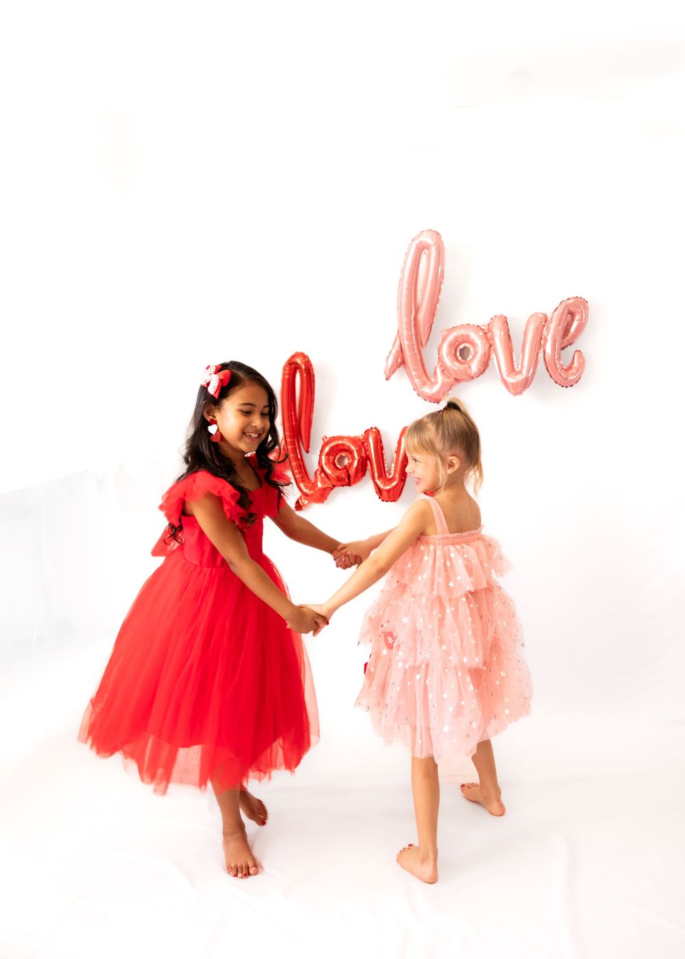 Two children in tulle dresses, one red and one pink, dance together with love balloons in the background.