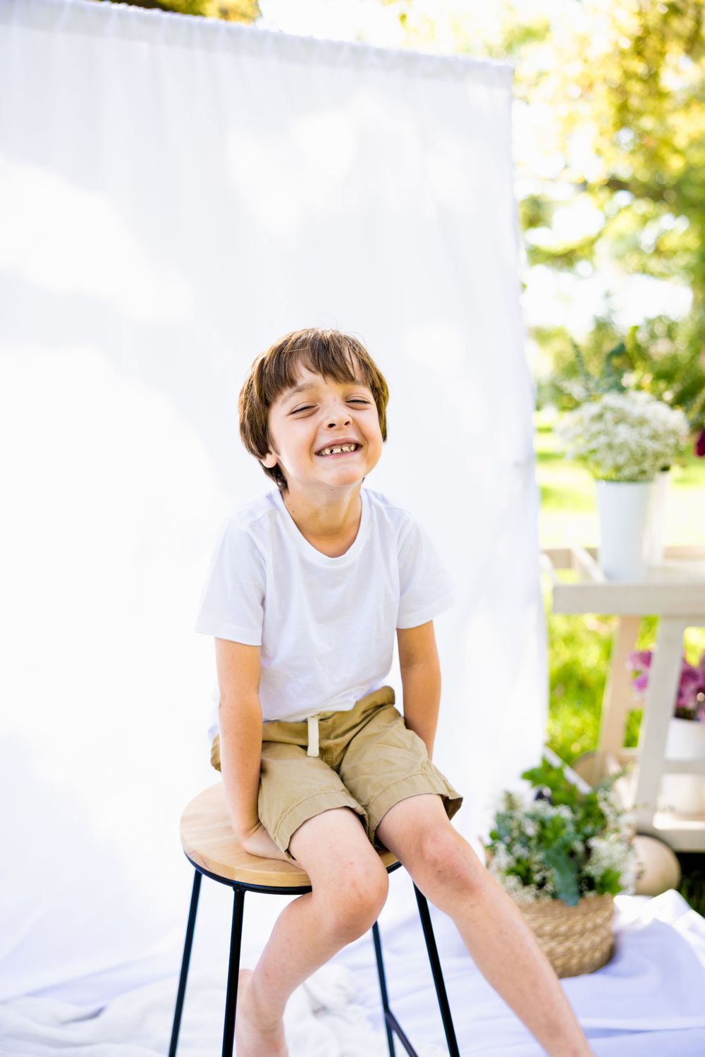 Young child in white t-shirt and khaki shorts sitting on wooden stool against white backdrop in sunny outdoor setting.