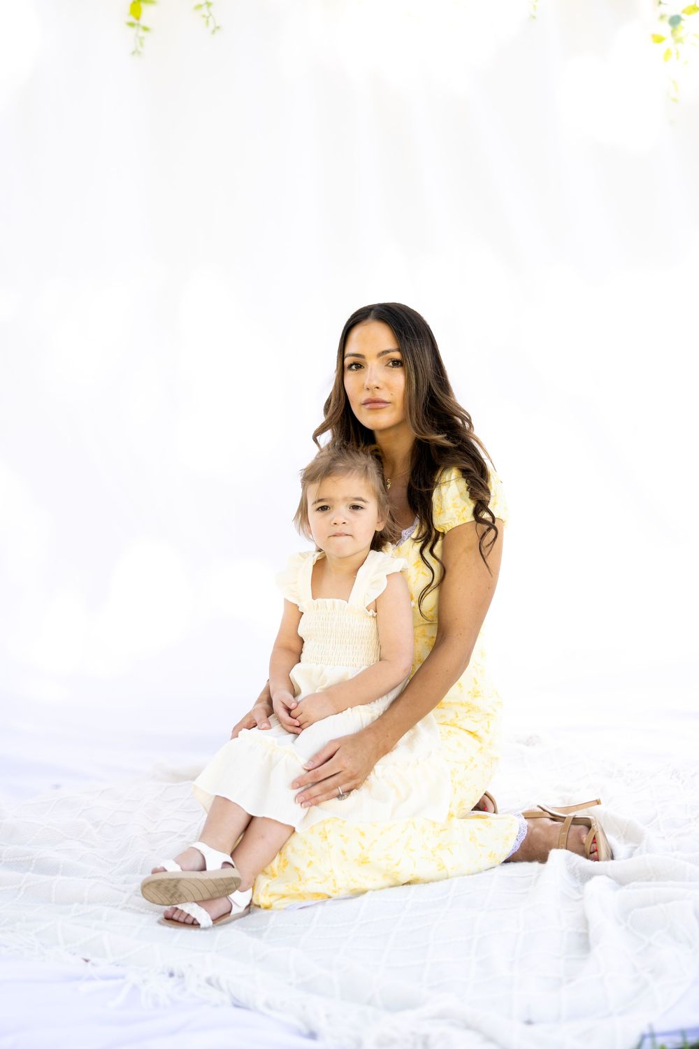 Two people wearing light colored outfits sit together on a white backdrop for a portrait session.