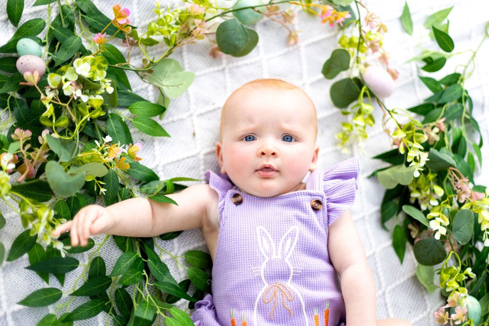 A small baby wearing a purple romper sits surrounded by green leafy vines and yellow flowers against a white backdrop.