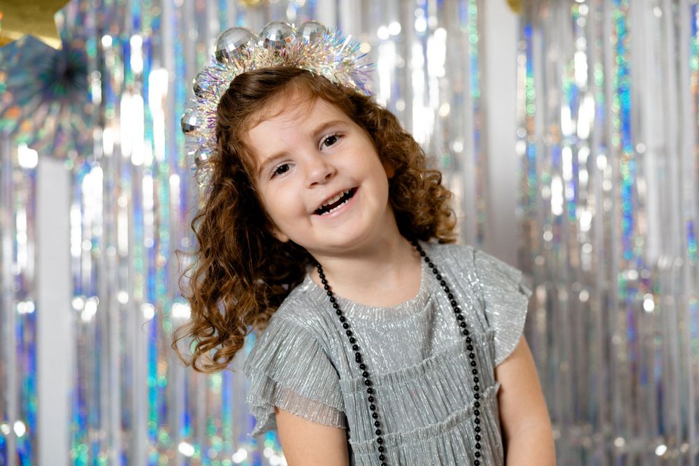 Child in silver dress and crown smiling against sparkly silver backdrop.