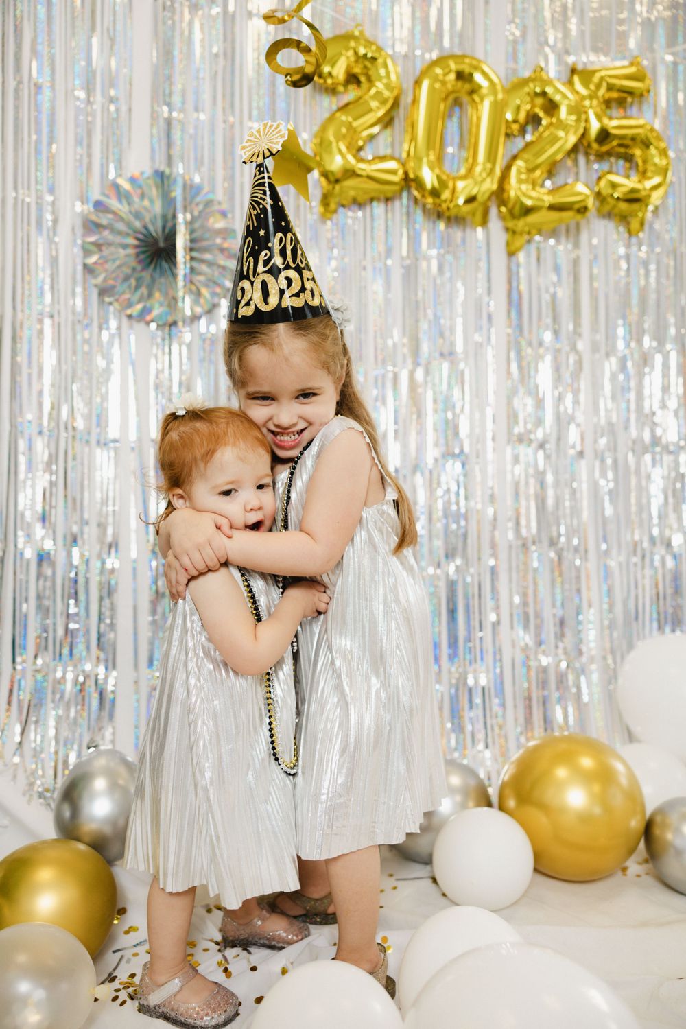 Two children in silver dresses hugging under 2025 gold balloon decorations.