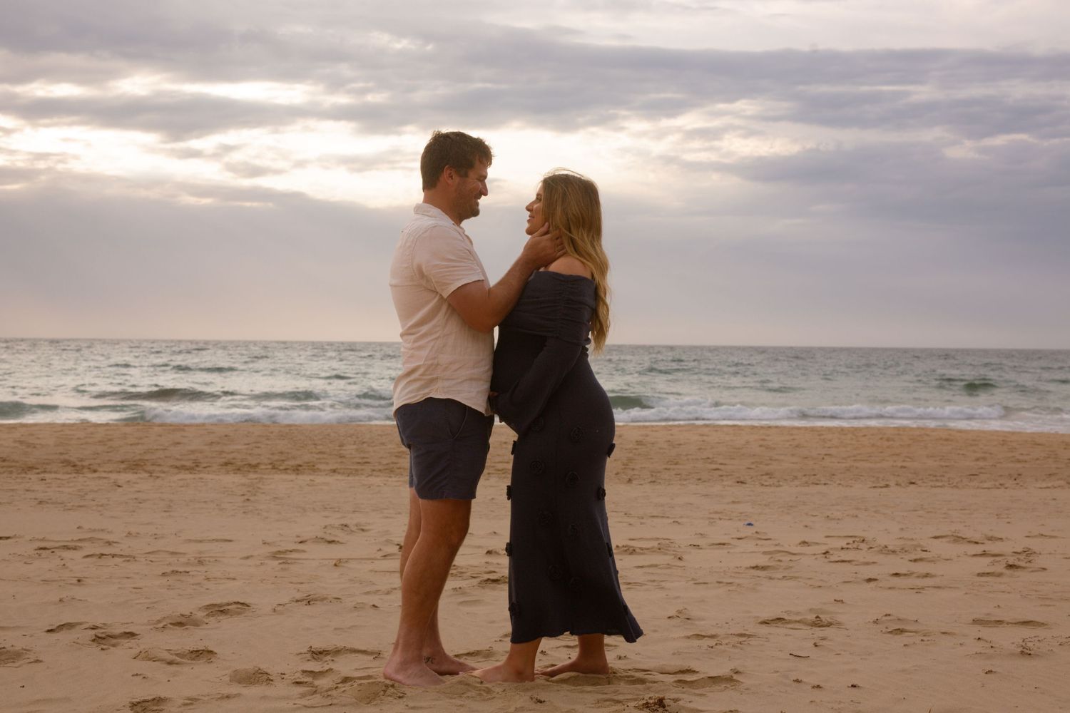 A couple shares an intimate moment at sunset on a beach with waves in the background.