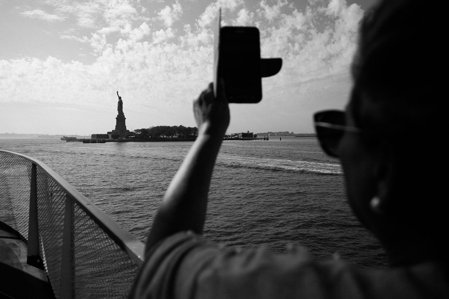 Silhouetted figure photographing Statue of Liberty from boat on cloudy day.