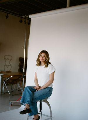 A person in a white t-shirt and blue jeans sits on a silver stool against a white backdrop in a studio setting.