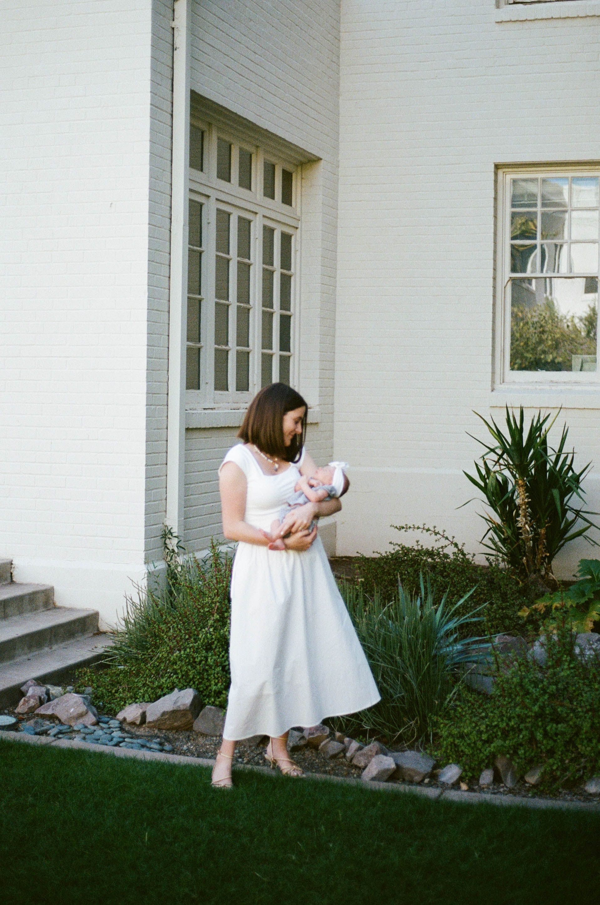 A woman in a white dress stands outside a traditional white house with a garden in the foreground.