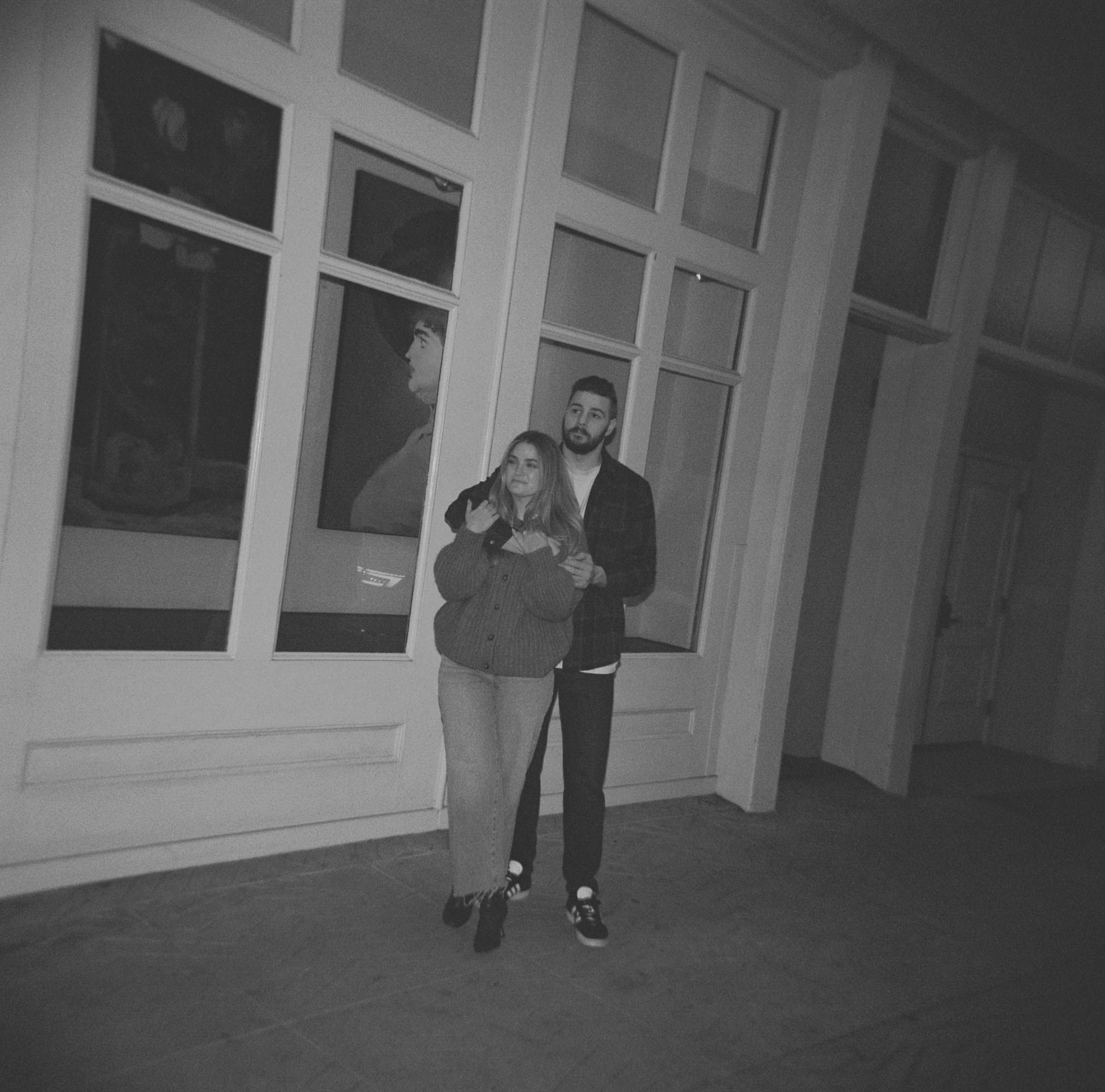 Black and white photo of two people standing in hallway with large windows.