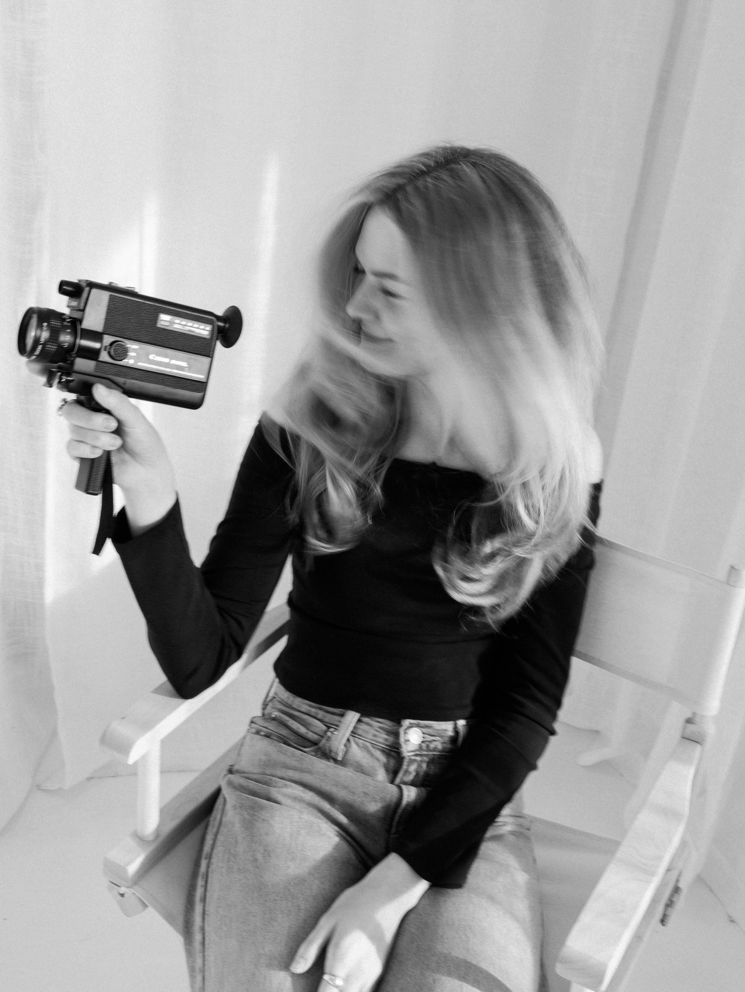 Black and white photo of someone holding a vintage video camera while sitting in jeans and black top.