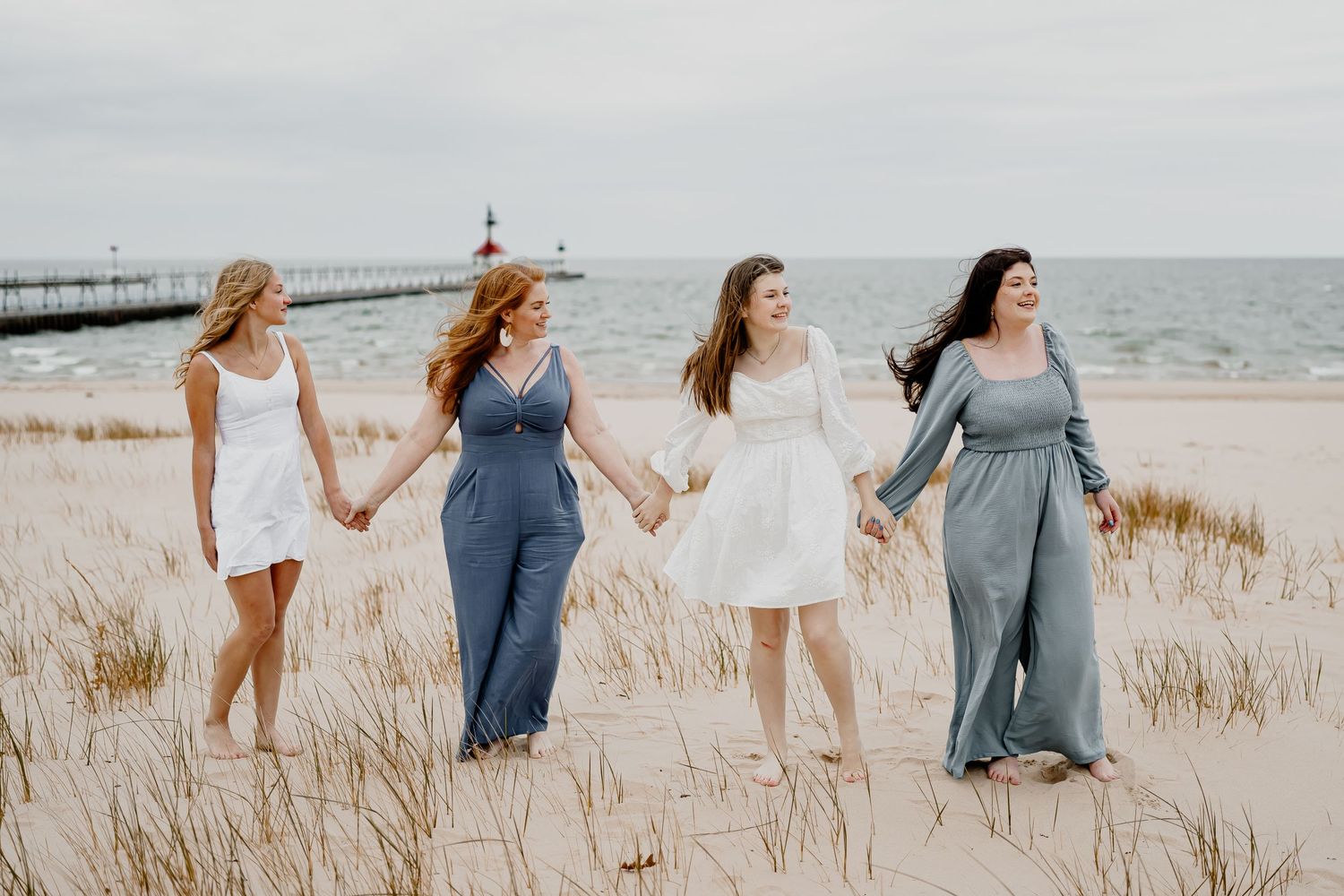 Four friends in flowing dresses walk hand in hand along a sandy beach path with tall grass and ocean views.